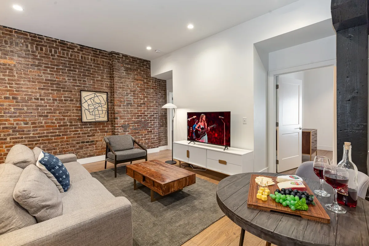 A stylish living room with an exposed brick wall, a large TV displaying a live performance, a comfortable sofa, and a coffee table arranged with snacks—inviting for group gatherings. The space blends urban charm with functionality, ideal for relaxing, watching movies, or celebrating with friends.