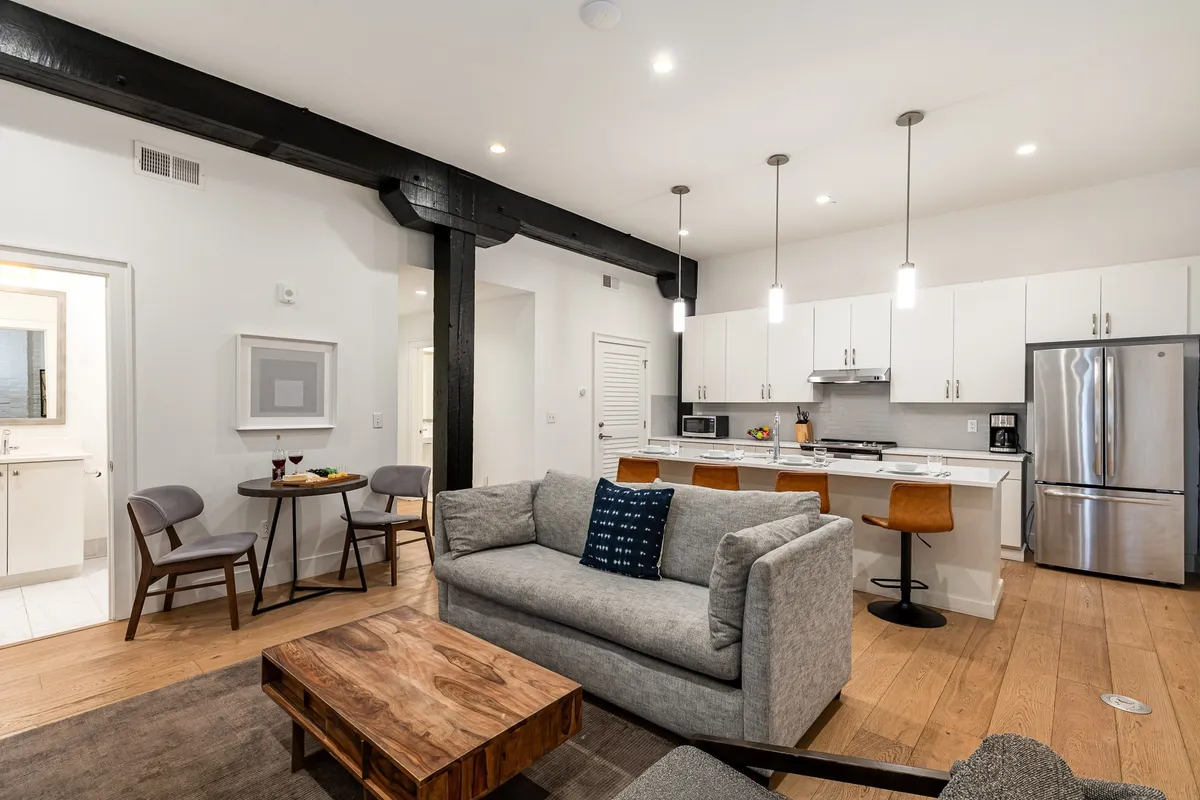 An open-concept living area showing a comfortable sofa, wooden coffee table, dining area, and kitchen island. Morning sunlight pours in, enhancing the inviting ambiance and showcasing group-friendly design.