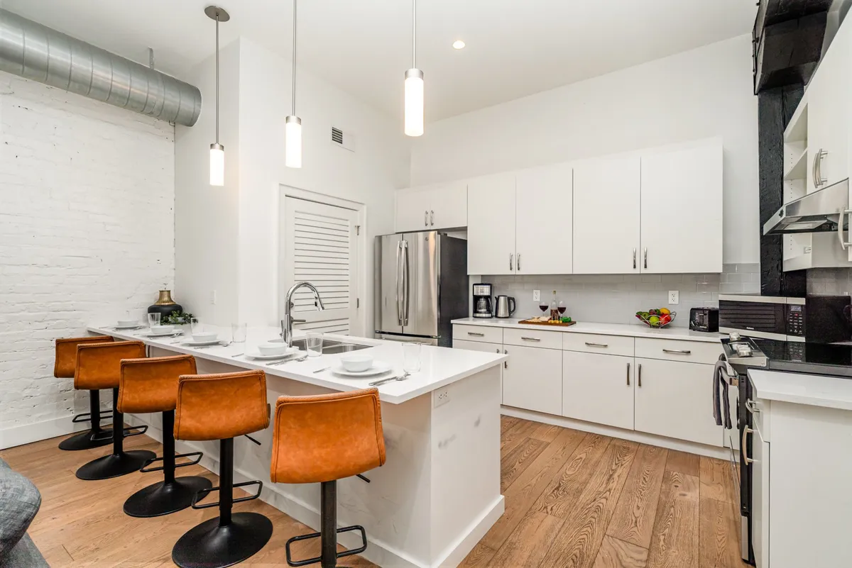 A bright, contemporary kitchen featuring a spacious island, several bar stools for group seating, white cabinetry, and modern appliances. The setup is designed for shared meals, cooking together, and easy flow between kitchen and living areas.