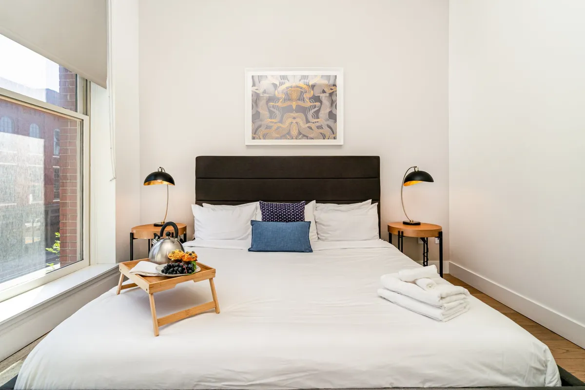 A cozy bedroom with a large bed dressed in white linens and colorful decorative pillows, complemented by artwork on the wall and a bedside lamp. A breakfast tray with food and a kettle sits atop the bed, hinting at leisurely mornings and comfort.
