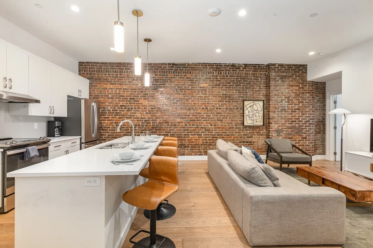 A spacious kitchen and living area with an exposed brick wall, a large kitchen island with multiple modern bar stools, gleaming stainless steel appliances, and a comfortable sofa and coffee table. The bar stools at the island invite guests to gather while someone cooks or prepares drinks, creating a lively and communal atmosphere.