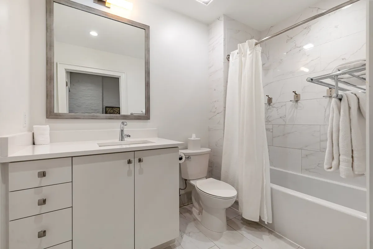 A calming bathroom with a bathtub fitted with a shower curtain, a wide mirror above the sink, and fresh towels neatly arranged. The minimalist decor and gentle lighting evoke a sense of relaxation and renewal.