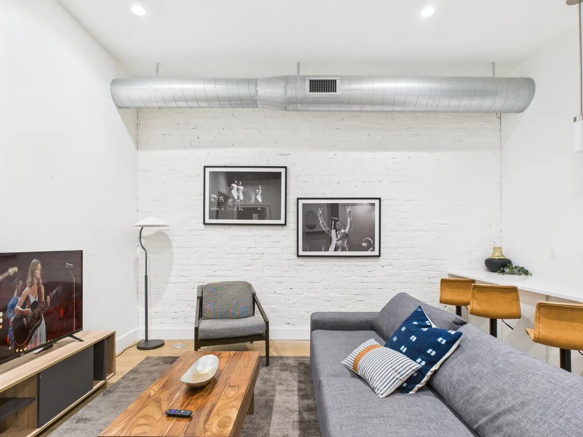 A modern living room featuring a cozy sofa, stylish coffee table, wall-mounted TV, and bar stools. Artwork adorns the walls, and the space feels inviting and vibrant—ideal for both small gatherings and quiet moments.