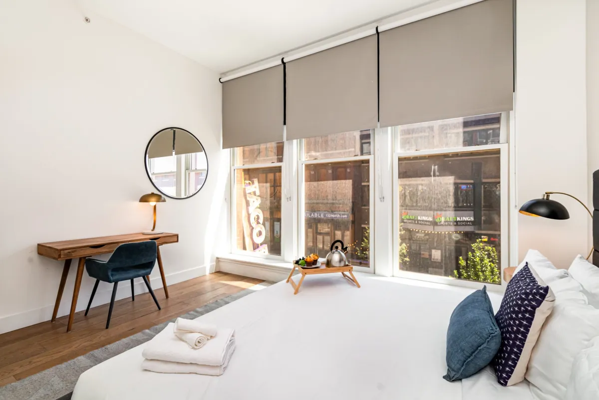 A tranquil bedroom with a plush bed topped with multiple pillows, a desk with a chair positioned near large windows, and a tray with a kettle for tea or coffee. The space is filled with natural light, and the overall feel is one of calm and relaxation, ideal for both rest and productivity.