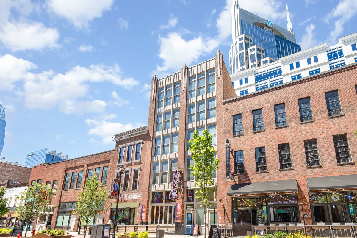 The image showcases the vibrant exterior of Nashville's historic Saddlery building surrounded by lush green trees, with views of lively outdoor seating areas, colorful signage, and the striking mix of classic brick architecture and modern skyscrapers. Locals and visitors are seen enjoying dining at outdoor cafes, exploring unique boutiques, and soaking up the energetic downtown atmosphere.