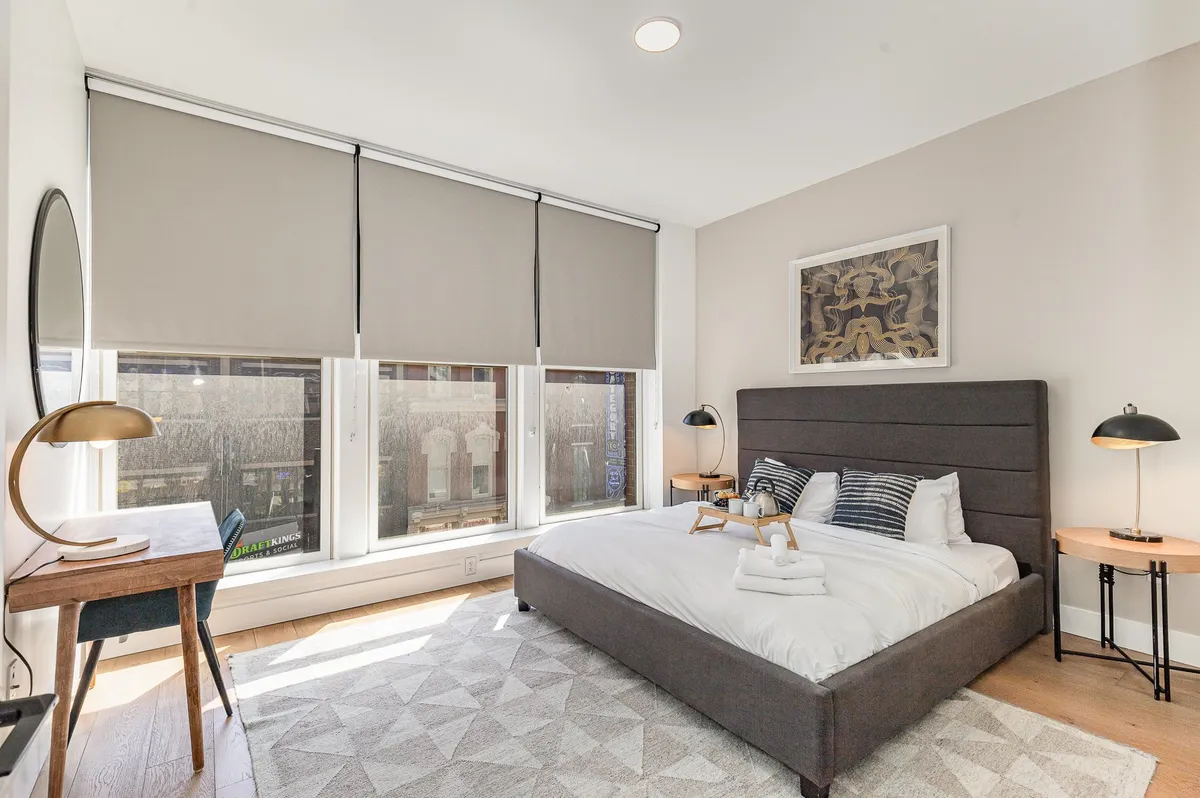 A peaceful bedroom with a comfortable bed, a window with shades letting in natural light, a desk with a chair, and artwork on the wall. The setting is tranquil and supportive of both relaxation and productivity.