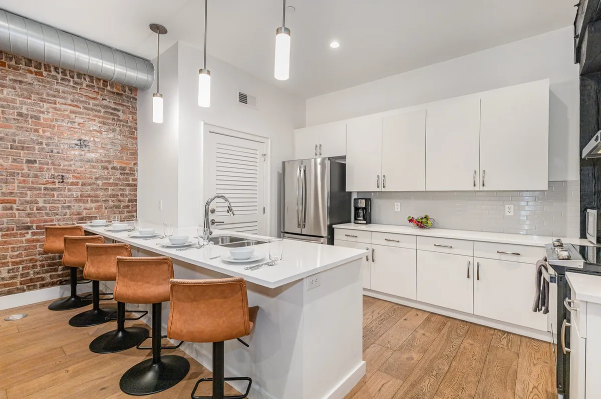 A modern kitchen with an exposed brick accent wall, sleek white cabinetry, stainless steel appliances, and a row of stylish bar stools. The kitchen looks ready for hosting, with plenty of counter space and a contemporary, inviting vibe.
