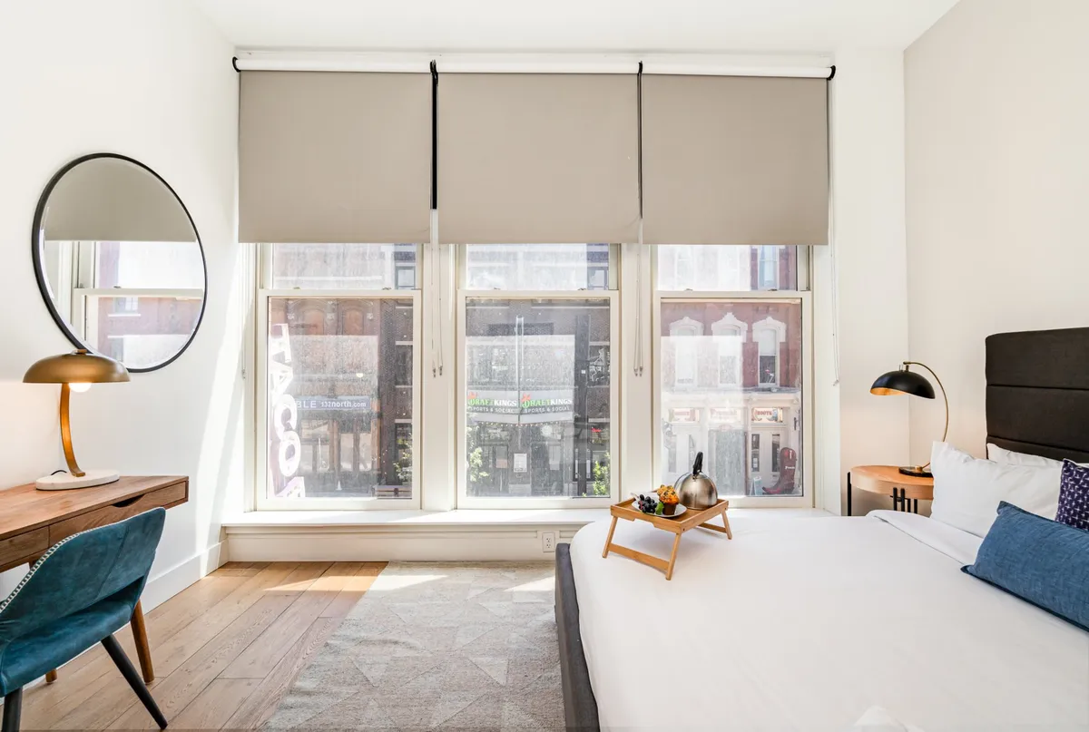 A stylish bedroom featuring a comfortable bed with decorative pillows, a desk and chair set by large windows offering a city view, and a fruit tray on the bed. The room blends comfort with a sense of place, connecting relaxation with opportunity for work or leisure.
